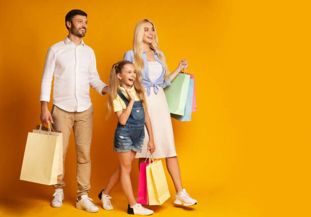 Family Shopping. Parents And Little Daughter Carrying Shopper Bags Walking In Studio Over Yellow Background. Empty Space